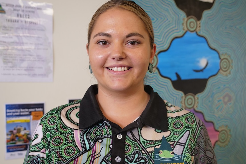 A young womanl with brown eyes, blonde and brown hair, wearing a colourful shirt smiles at the camera.