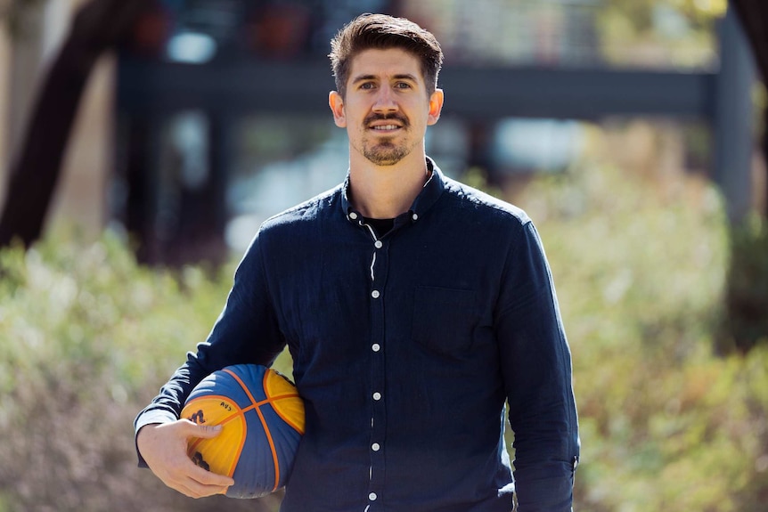 A tall man wearing a navy blue shirt holds a basketball and stands in a park