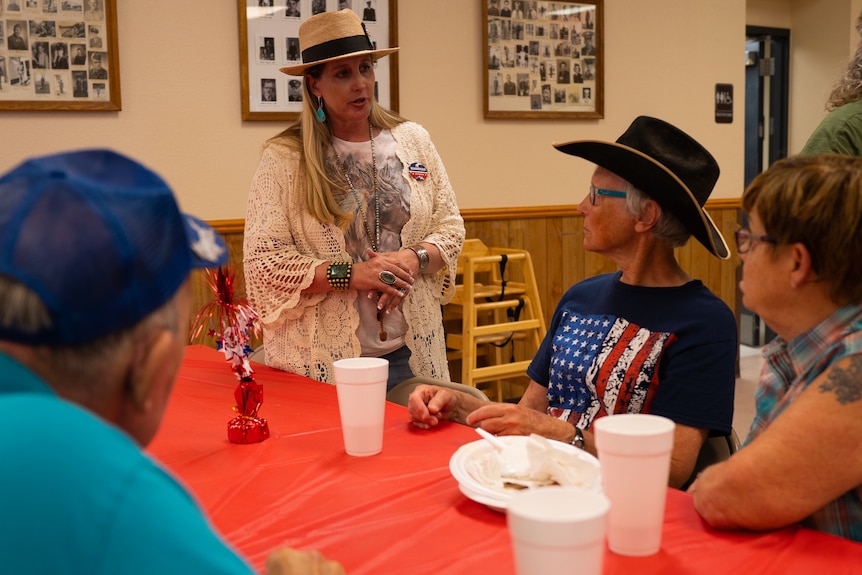 Woman stands at table talking with constituents. 