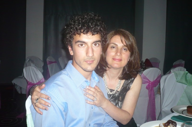 A young man sitting at a table at a family function with his mother with her arms around him.