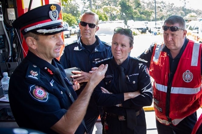 Female firefighter stands with arms crossed listening to a male firefighter talk, other firefighters are nearby also listening.