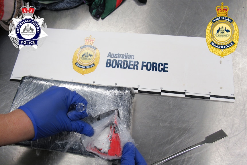 Hands in blue gloves hold a knife that has been used to cut open a packaged wrapped in plastic, revealing a white substance.