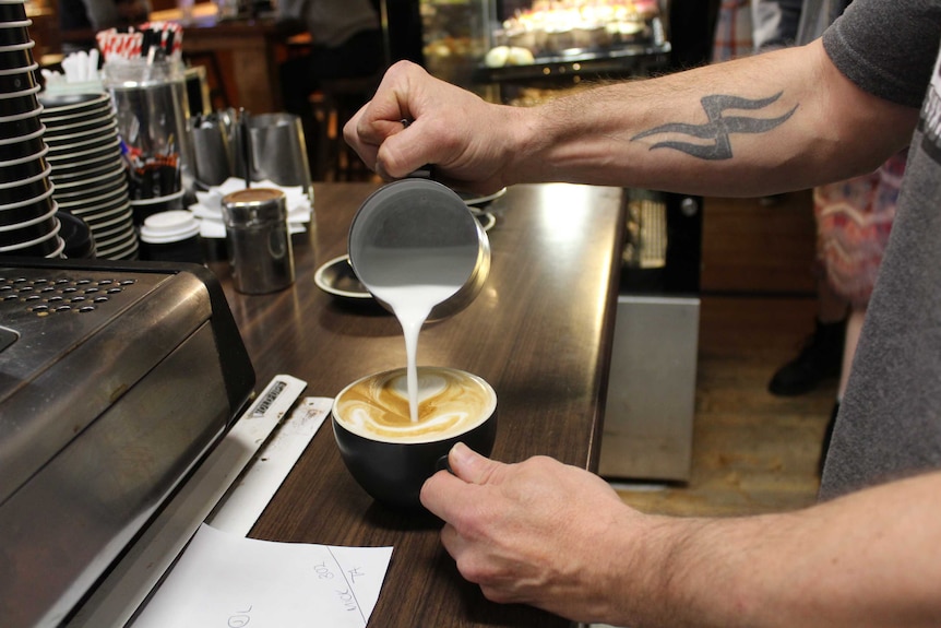 Close-up of Mark pouring milk from a metal jug into a cup of coffee.