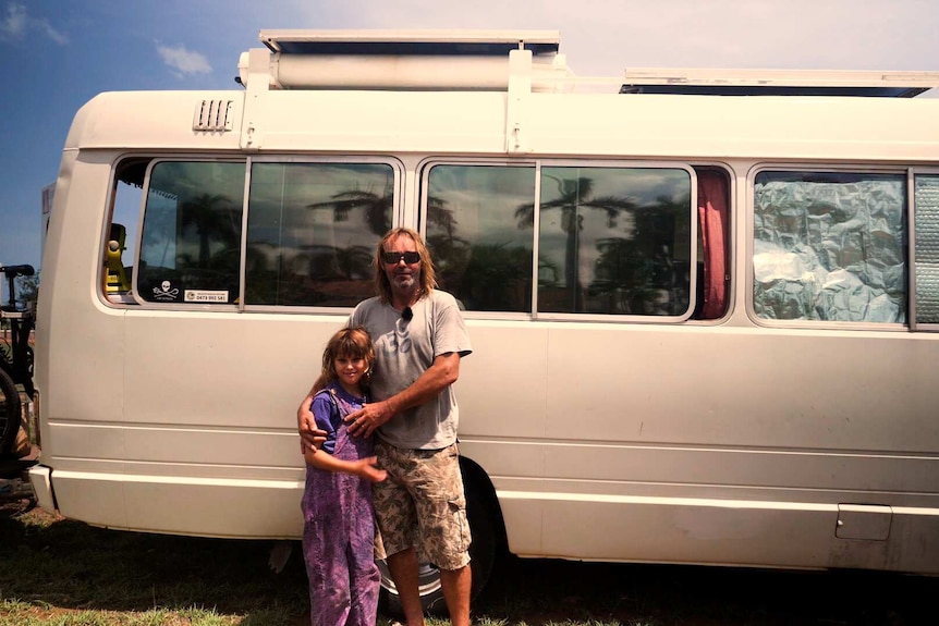 Man and daughter standing outside on drivers side of white coaster bus. Sunny day.