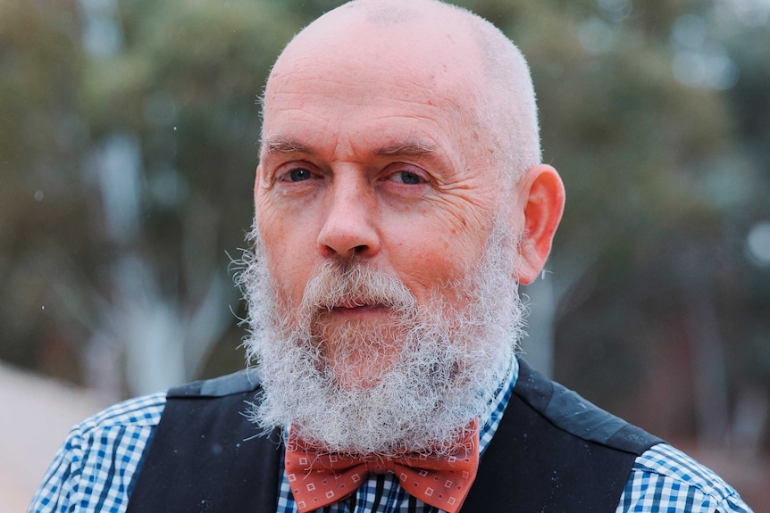 A bald man with a white beard is looking at the camera, wearing a bow-tie.