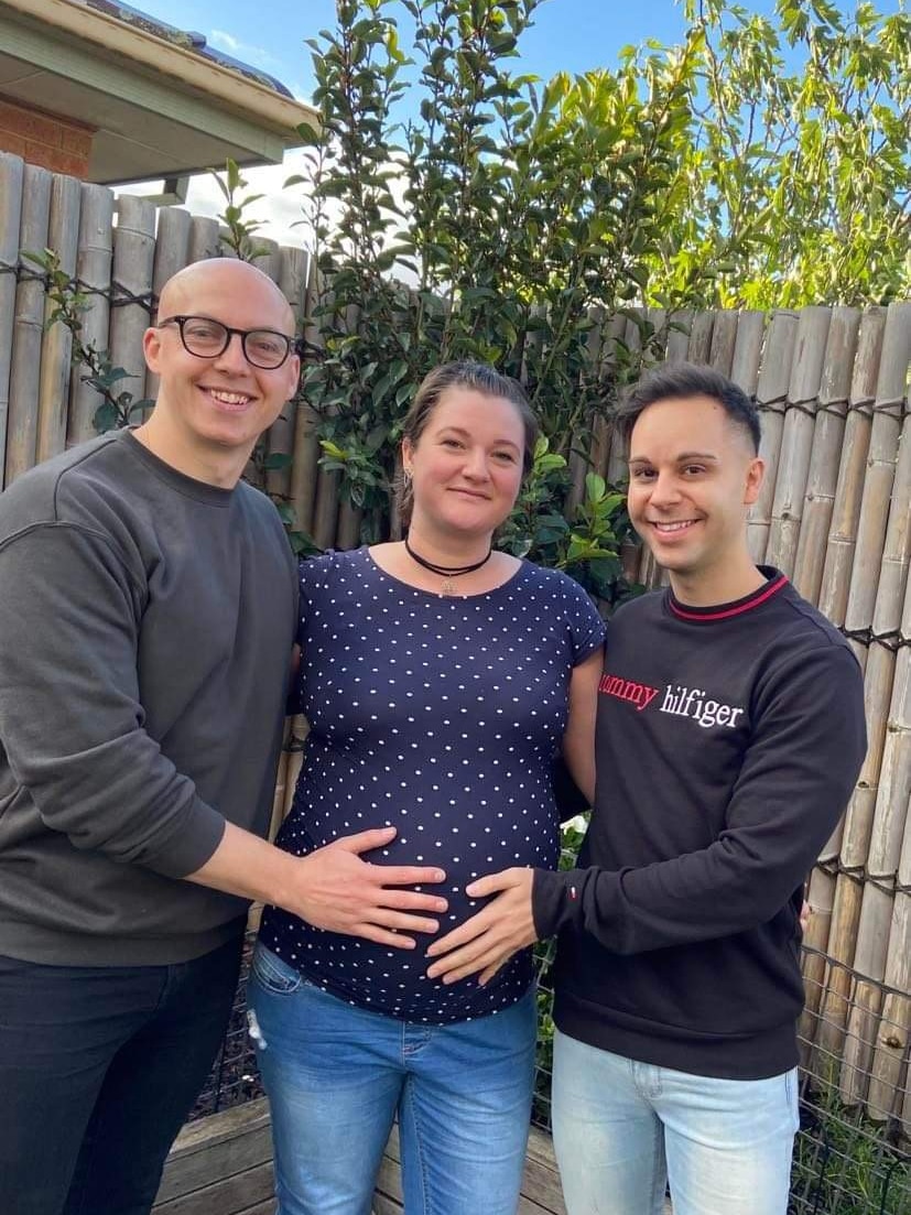A pregnant woman stands between two men who are resting their hands on her bump. They smile and stand in a plant-filled garden..