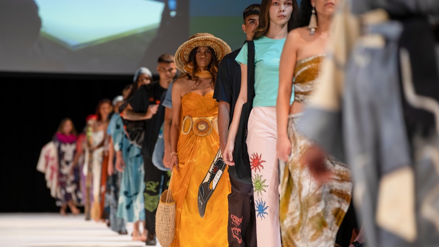 A line of fashion models walk in single file on a catwalk. 