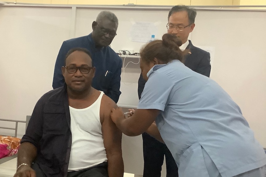 Seated man with shirt half undone receives vaccination from lady in blue uniform, while two men look on behind.