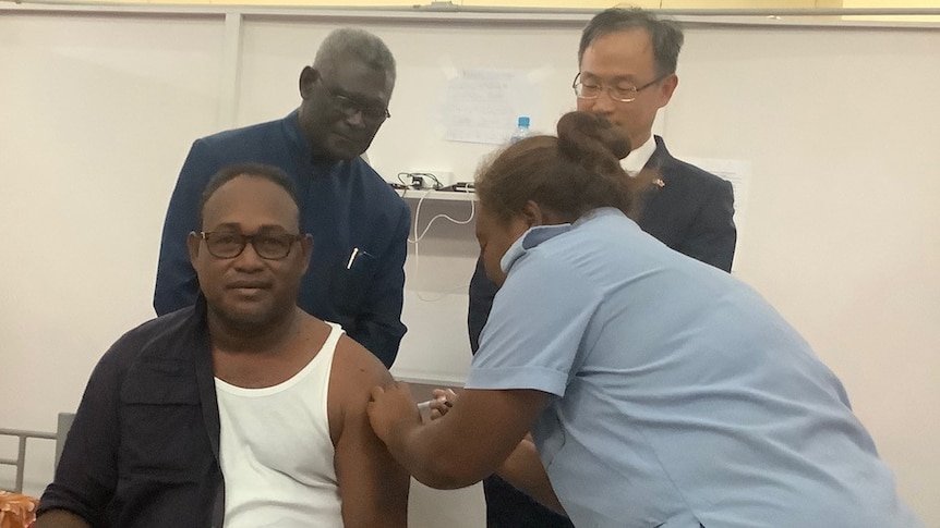 Seated man with shirt half undone receives vaccination from lady in blue uniform, while two men look on behind.