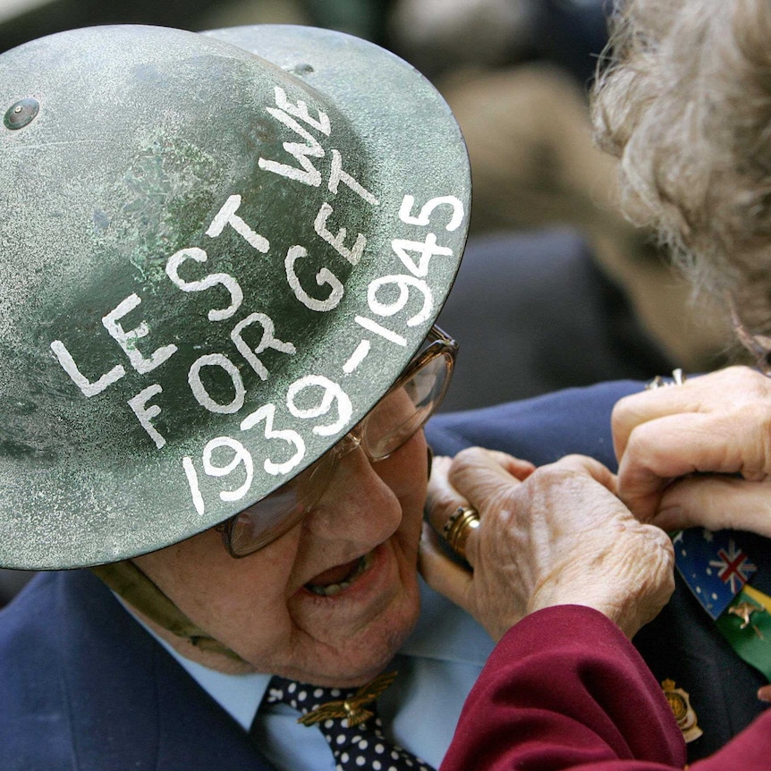 senior man wearing helmet with the words "lest we forget 1939 to 1945". Senior woman pins australian flag badge to his suit.