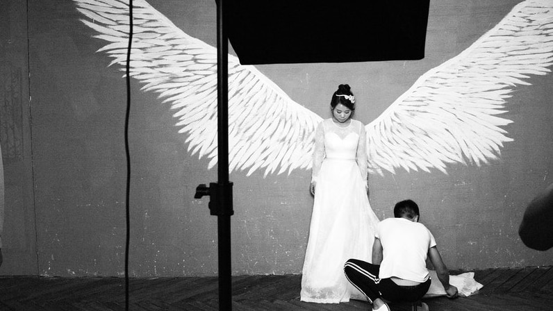 A bride poses for a prewedding photo shoot. She stands against a wall, in between a set of angle wings as a man fixes her dress.