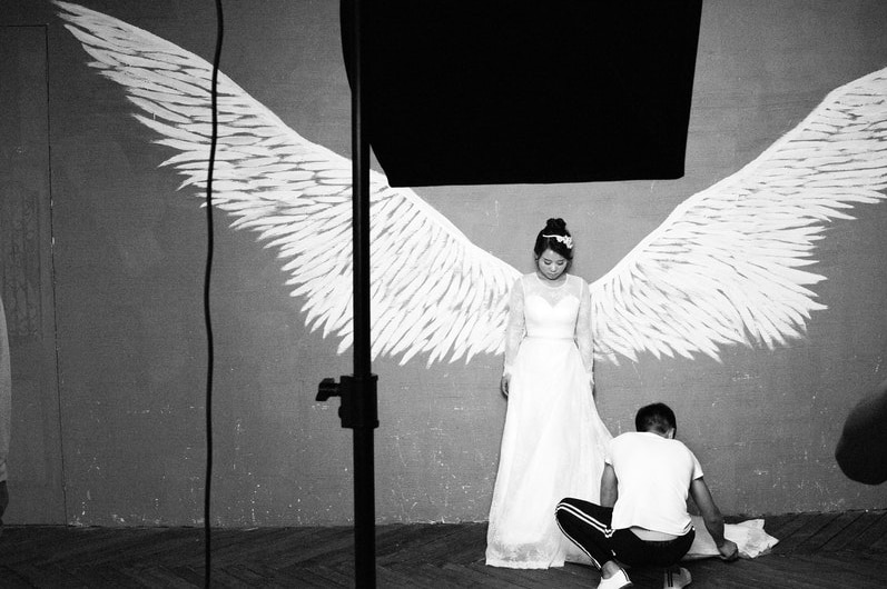 A bride poses for a prewedding photo shoot. She stands against a wall, in between a set of angle wings as a man fixes her dress.