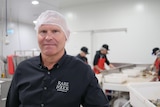 A mean wearing a dark shirt and white hair net stands in a factory and looks into the camera.