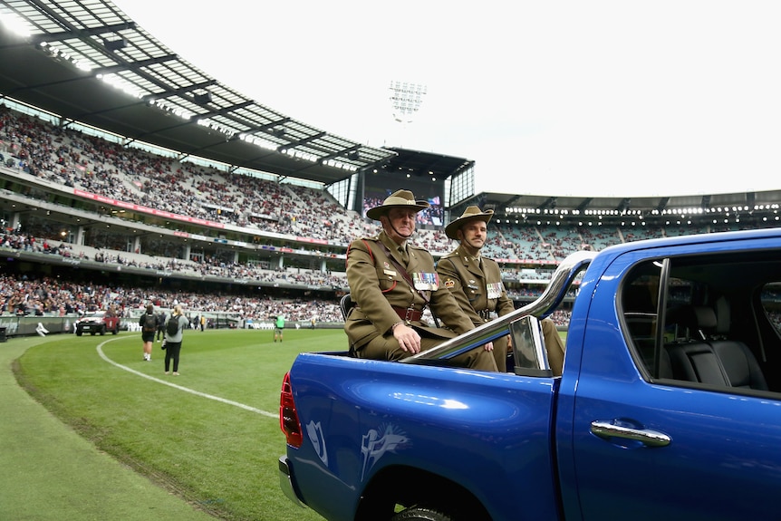 Two men in military clothes sit in the back of a ute at the Anzac Day AFL game between Essendon and Collingwood.