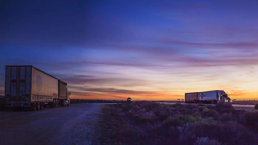 The sun rises over a truck, left, sitting in a truck-stop as trucks speed past on the highway, right.