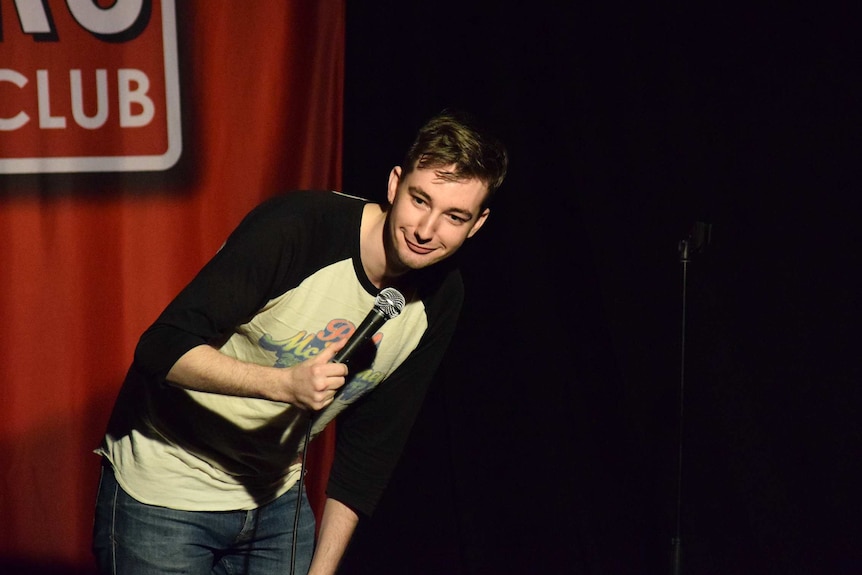 Comedian David Rose on stage at a comedy gig, he is holding a microphone, leaning forward and grinning.