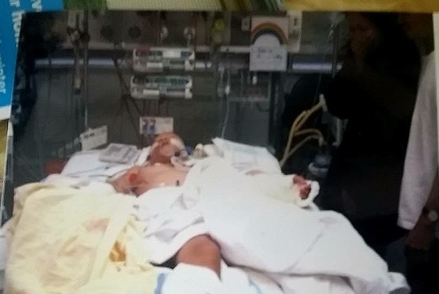 A little boy lies unconscious on a hospital bed covered in blankets with tubes plugged into him.