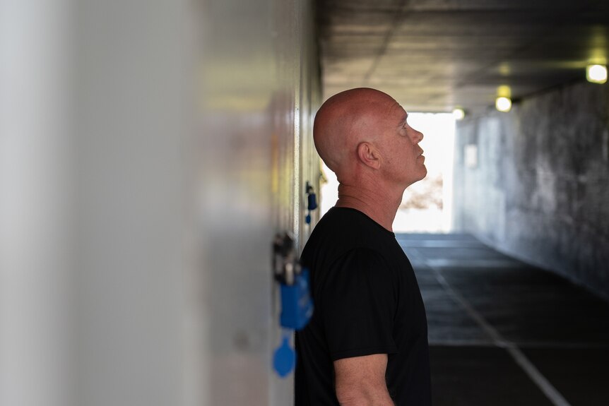 A bald man in a black t-shirt stands side on leaning against a wall