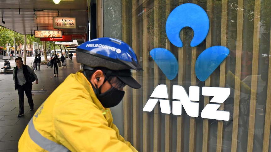 bicycle courier wearing protective mask passes ANZ logo in city street