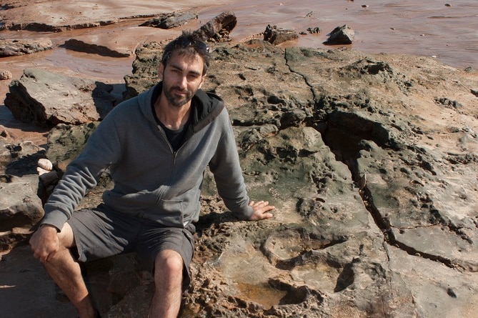 A man with dark hair and beard sits on a big rock on a beach.