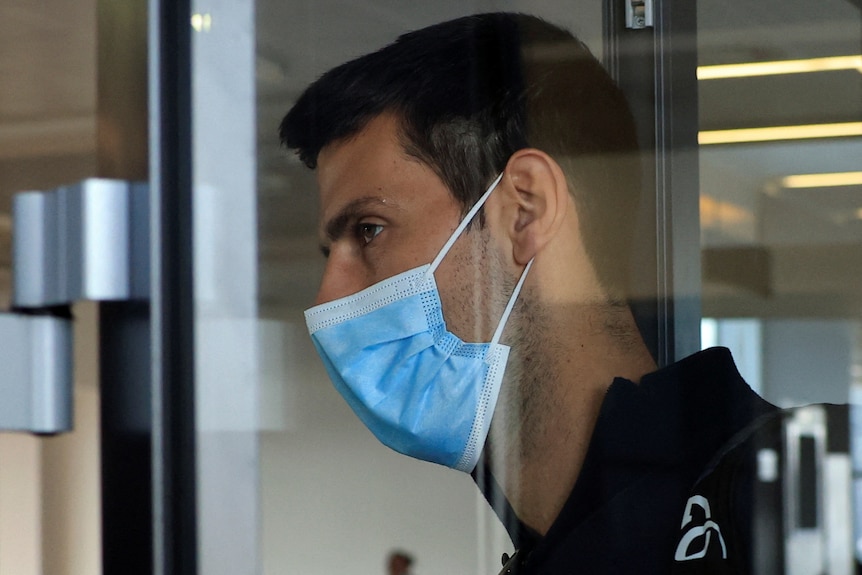 Novak Djokovic, wearing a mask, is seen through glass at Belgrade airport.