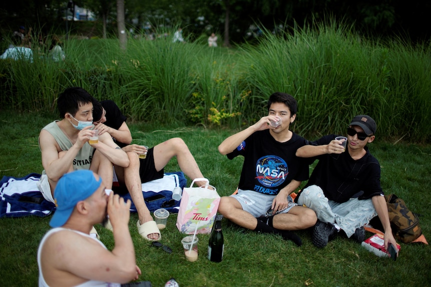A group of young Chinese people sit in a park with a picnic spread in the middle 