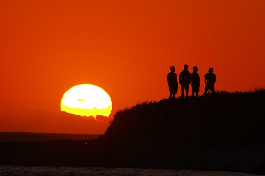 Four figures in silhouette standing on headland watching sunset amid red sky