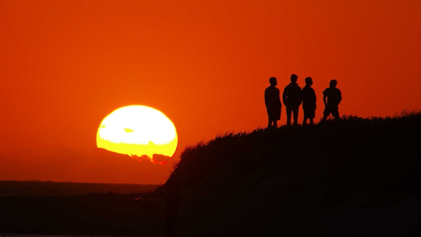 Four figures in silhouette standing on headland watching sunset amid red sky