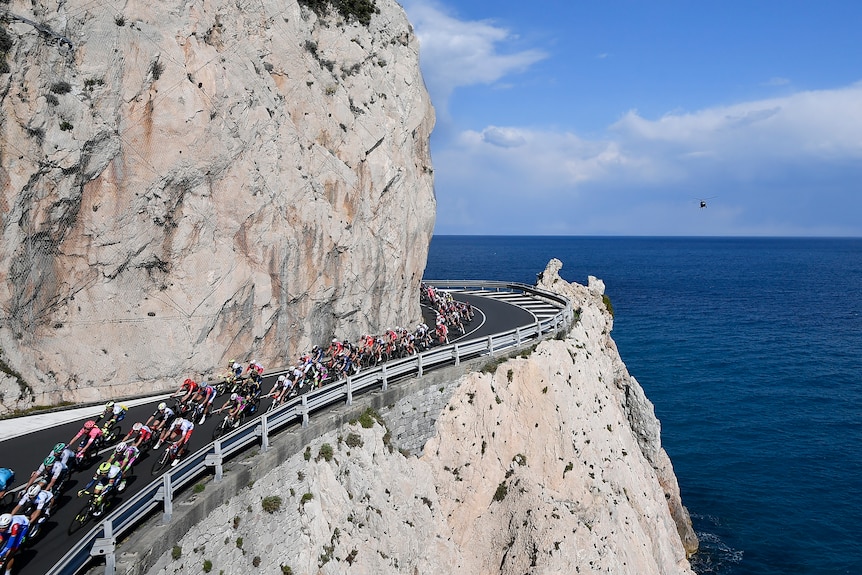Cyclists can be seen riding on a road alongside steep white cliffs and the sea