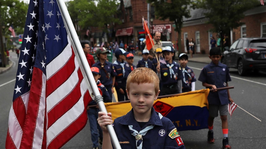 A young boy holding an American flag leads a group of Boy Scouts in a parade.