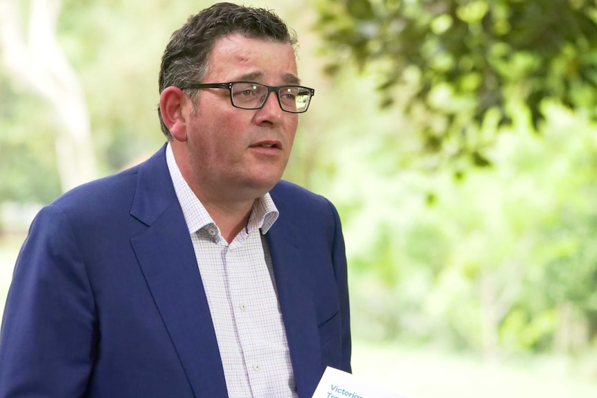 Victorian Premier Daniel Andrews wearing a blue suit jacket and talking in a green leafy location.