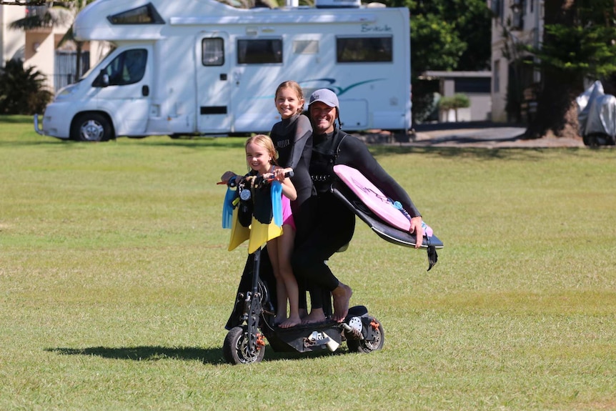 A man in a wetsuit sitting on a scooter, with two young girls in front of him, carrying bodyboards under his arm.