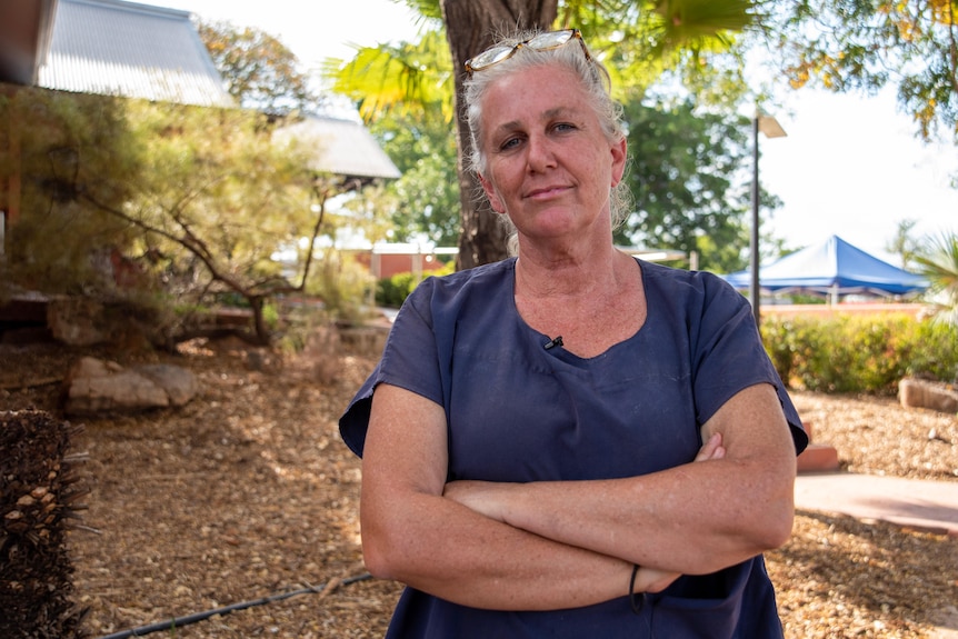 A woman with gray hair stands in a leafy yard with her arms folded.