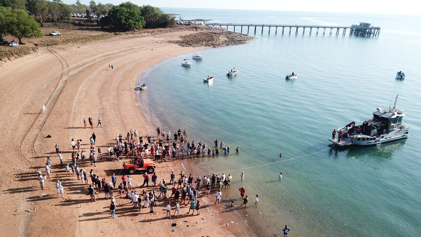 A drone photo of a group of people gathered around a car entering the water. It is daylight.