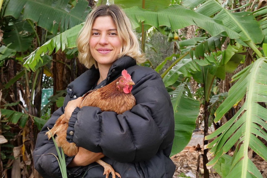 A woman in a puffer jacket holds a chicken in her arms, standing in a banana plantation.