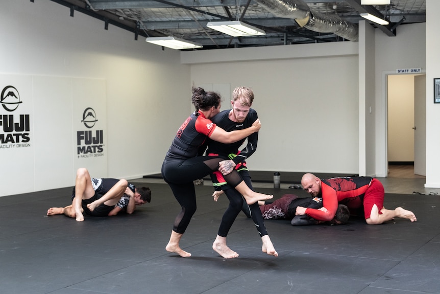 A man and woman stand up while grappling on a mat in a gym.