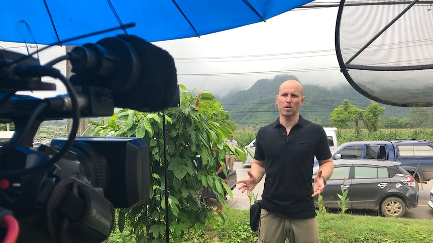 Liam Cochrane standing in a green field with mountains and cars behind him, speaking towards a video camera.