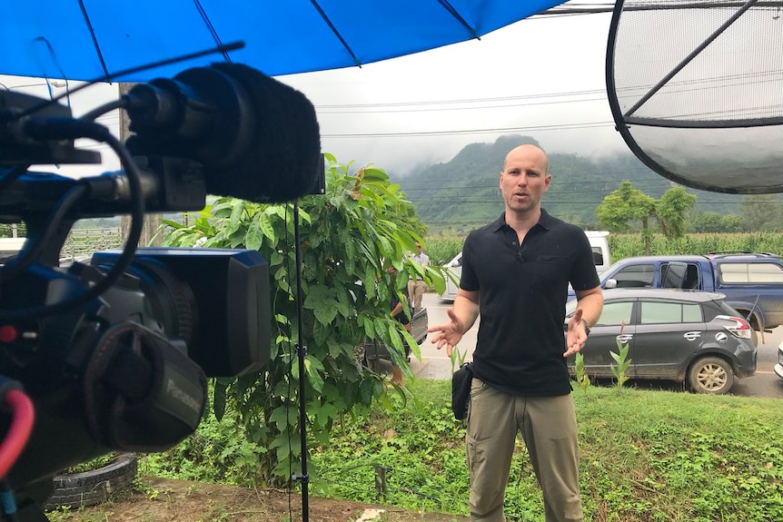 Liam Cochrane standing in a green field with mountains and cars behind him, speaking towards a video camera.