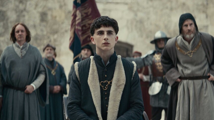 The actor Timothée Chalamet as King Hentry in formal wear and bowl cut, seated with men in the background