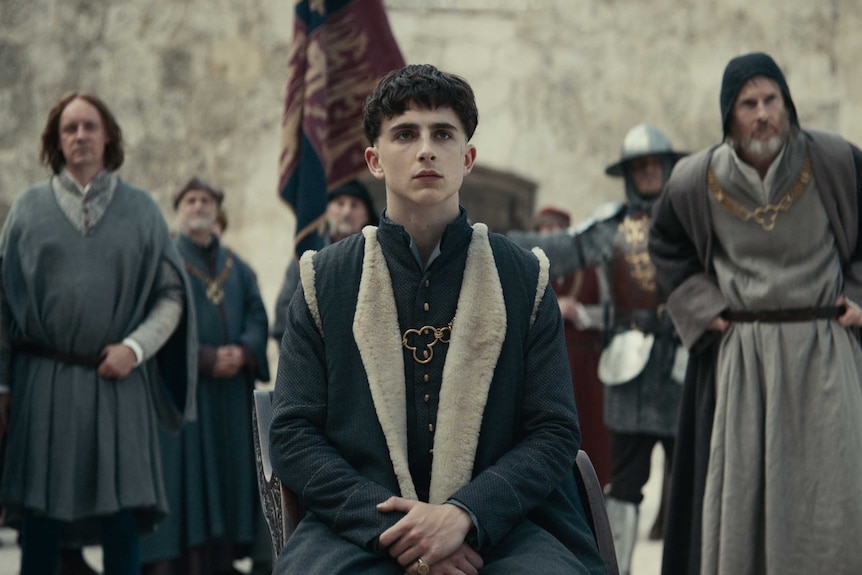 The actor Timothée Chalamet as King Hentry in formal wear and bowl cut, seated with men in the background