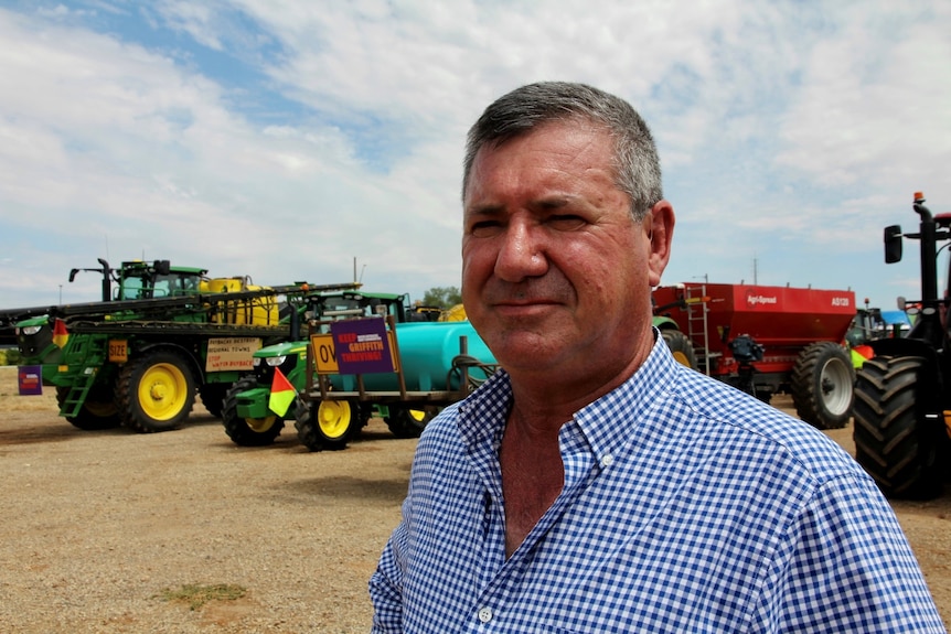 A man looks at the camera with a blank expression. He is standing in front of farm machinery.