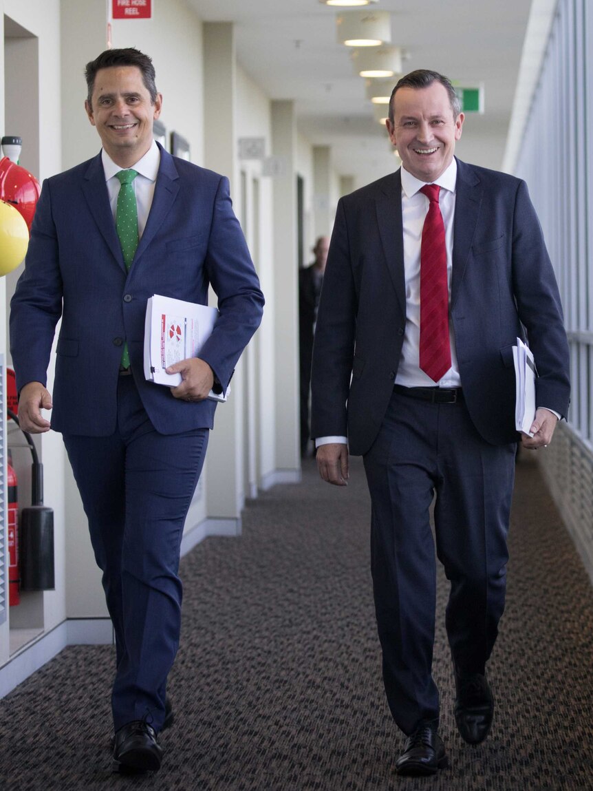 Two men in suits walk down a white corridor.