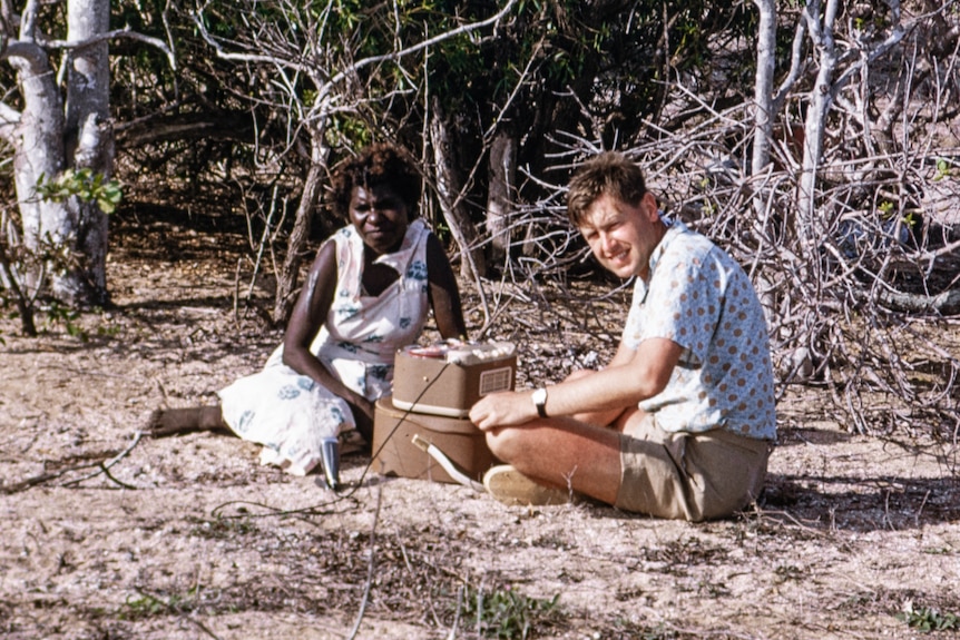 A white man and an Indigenous woman sit on the ground with a recording device between them.