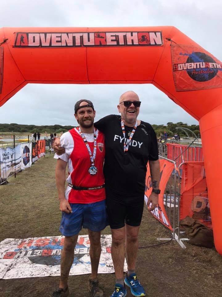 An older man with glasses embraces his son who is in his twenties at the finish line of a race.