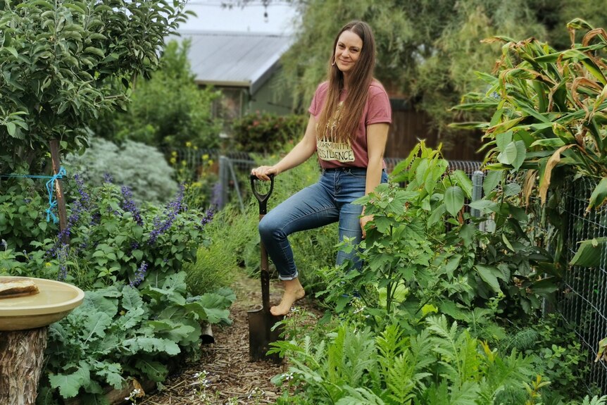 A woman with long brown hair, wearing blue jeans and a T-shirt, stands in a leafy garden, with her foot on a spade.