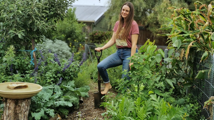 A woman with long brown hair, wearing blue jeans and a T-shirt, stands in a leafy garden, with her foot on a spade.