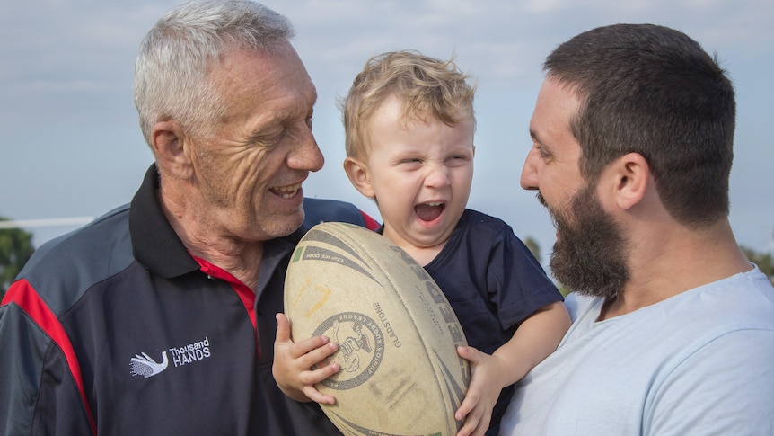 A a man with grey hair and a man with short hair hold a young boy who is laughing and holding a rugby league football.