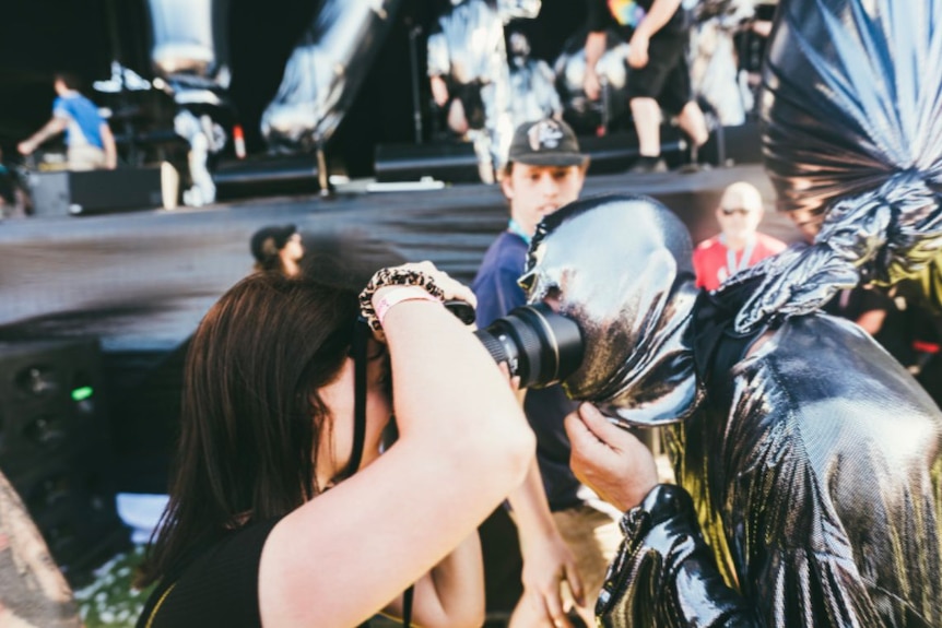 A photographer holds up their camera as a performer from TISM in a silver costume leans right into the lens
