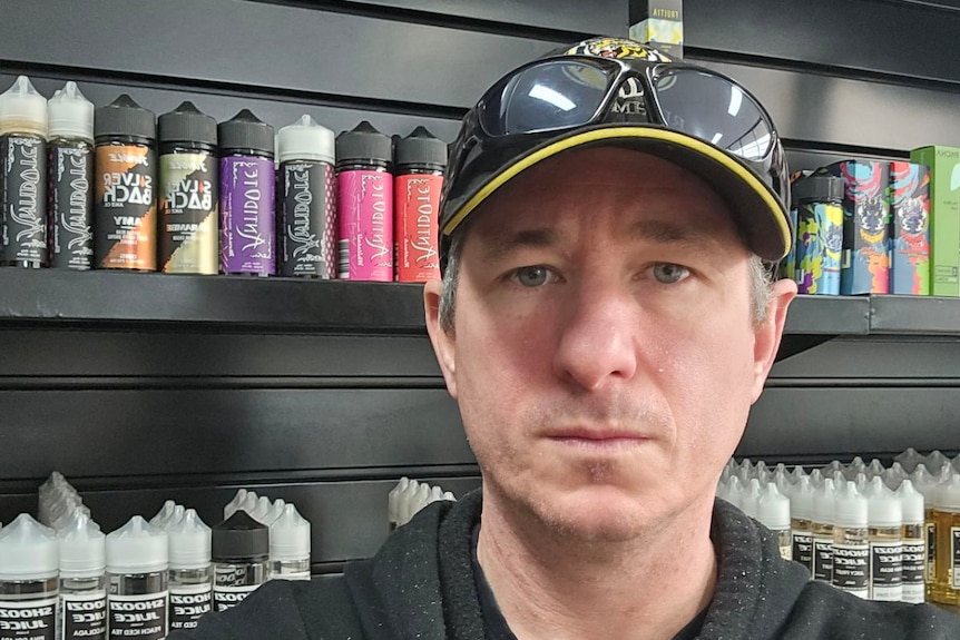A man looks serious, standing in front of shelves stocked with vapes.