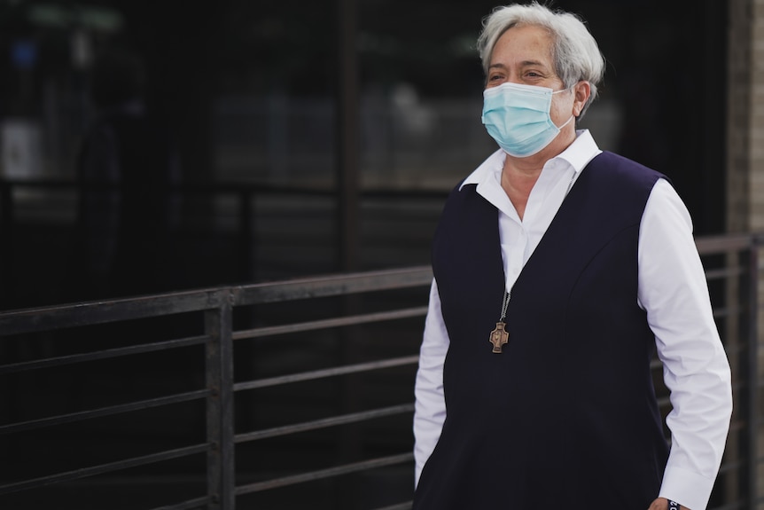 A woman with short hair wears a mask and a vest.